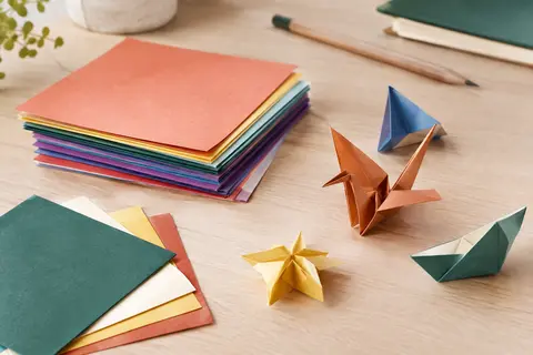 Feuilles carrées d’origami aux couleurs vives, empilées et pliées sur une table en bois clair.
